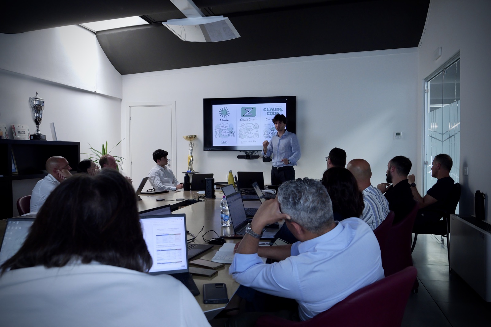 Riccardo Belli tiene un workshop attorno al tavolo riunioni, sullo schermo lo schema disegnato a mano di Claude e Claude Code, i partecipanti seguono dai laptop.